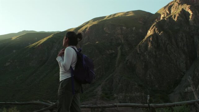 A female traveler walks and gazes at the immense mountains of the Peruvian Andes against a beautiful sunset.
