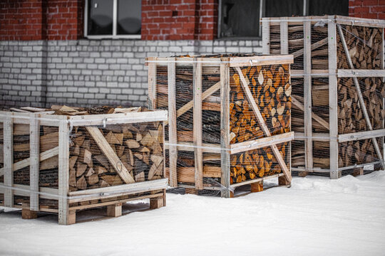 View of wooden crates filled with chopped firewood wrapped in plastic netting sitting on snow in front of a brick building in Latvia.