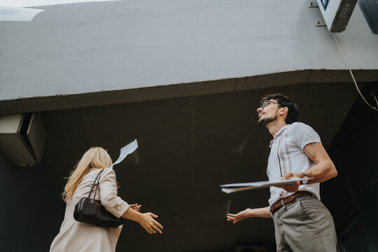 Two business colleagues stand under an overhang while loose papers fly from their hands. The pair react to the wind as documents scatter in a city entrance.