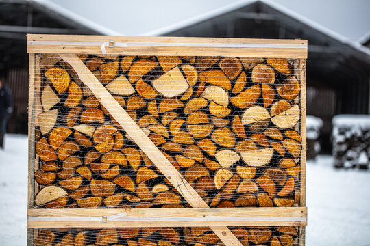 View of a wooden crate filled with stacked firewood wrapped in plastic netting in a snowy outdoor setting in Latvia.