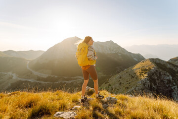 Obraz premium Female traveler with a yellow backpack stands on a mountain trail in the glow of the sunset. Woman with a hiking backpack enjoys the mountain scenery. Concept of adventure, freedom, and nature.
