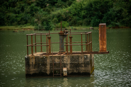 View of a weathered metal sluice gate structure in a calm lake with lush green vegetation in the background, Jos North, Plateau, Nigeria.