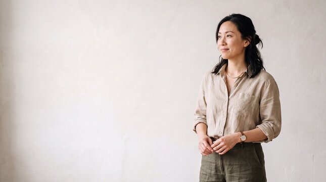 Entrepreneur standing by textured studio wall thinking about strategic plans. Asian female executive contemplating corporate decision making in modern office workspace.
