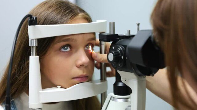 Young girl receives an eye exam using a slit lamp, with a light beam illuminating her eye and medical equipment in the background