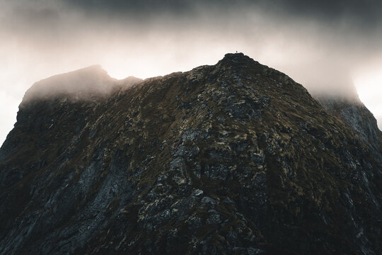 View of the rocky peak of Reinebringen mountain with mossy slopes and dramatic clouds under moody lighting in Moskenes, Nordland, Norway.