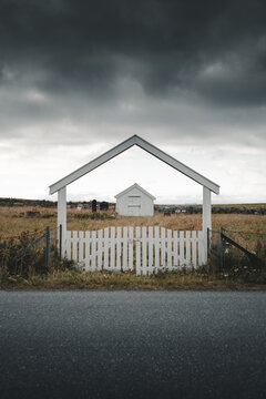 View of the white wooden gate framing the historic Flakstad Church in a grassy field under a dramatic cloudy sky in Ramberg, Nordland, Norway.