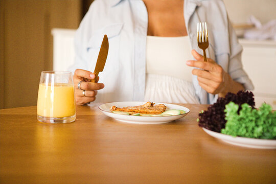 Woman holding knife and fork prepares to eat a healthy meal of grilled chicken and salad at a wooden dining table with a glass of juice nearby