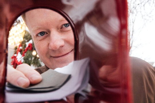 Man with light brown hair smiling while placing letters into a red mailbox during the holiday season, with festive decorations visible in the background