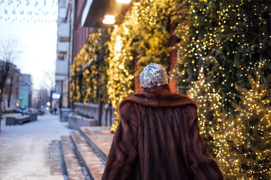 Woman in a brown fur coat walks along a snowy street adorned with festive lights and greenery, creating a winter holiday atmosphere in an urban setting