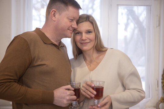 Middle-aged couple holding glasses of tea while standing close together in a bright room with large windows and soft natural light illuminating their faces
