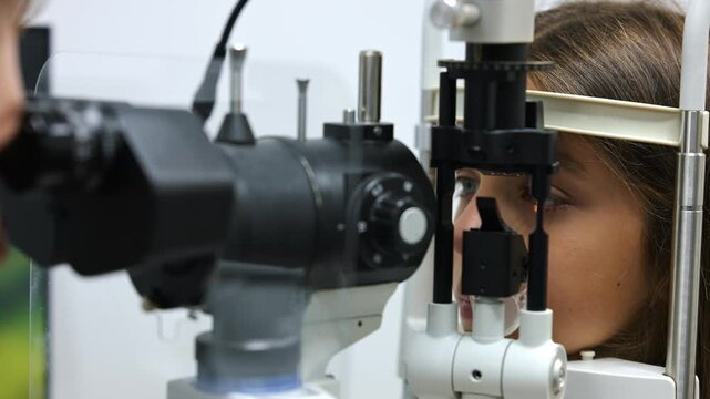 Young girl receives an eye exam using a slit lamp, with a light beam illuminating her eye and medical equipment in the background