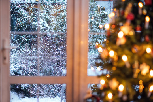 Snow-covered garden visible through a window with a decorated Christmas tree in the foreground, warm lights glowing softly, creating a cozy winter atmosphere with copy space