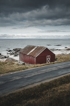 View of a traditional red wooden cabin at Vikten Beach with a rusty roof and white door frame, situated by the rocky coastline and dark ocean in Vikten, Nordland, Norway.