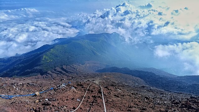 High-angle landscape shot taken from a steep, rocky volcanic trail looking down at a dramatic sea of white clouds stretching toward the horizon. The foreground features safety climbing ropes laid alon