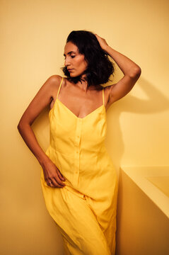 Elegant woman with dark hair wearing a yellow dress poses against a soft yellow background, showcasing a relaxed and stylish demeanor in a minimalist setting