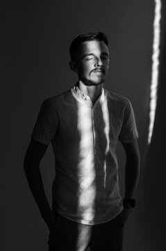 Black and white portrait of young man standing with eyes closed enjoying warm sunlight indoors. Handsome guy in casual shirt relaxed with contrast light beam pattern on face and body against wall.