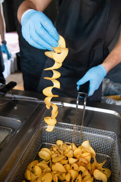 Food vendor wearing blue gloves lifts freshly fried spiral potato chips from a deep fryer during professional kitchen preparation.