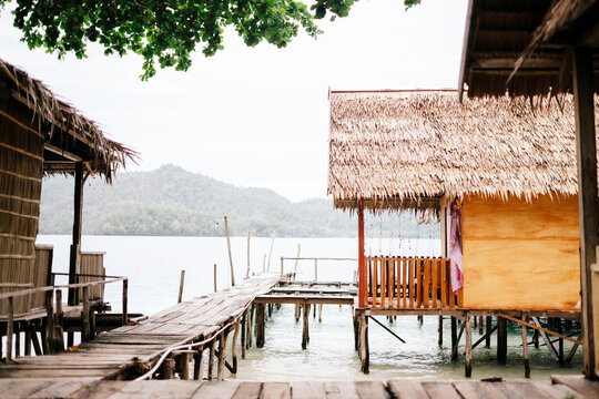 View of traditional wooden stilt houses with thatched roofs connected by a wooden walkway over clear turquoise water in Raja Ampat island, Southwest Papua, Indonesia.