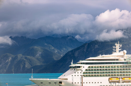 View of a large white cruise ship docked in front of dramatic, cloud-covered mountains and turquoise water in Haines, Alaska, United States.