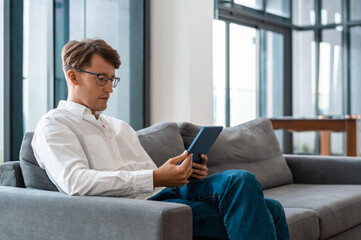 A professional man reads a tablet on a sofa in a bright modern office, photographic style, conveying remote work, focus, and productivity © ImageFlow