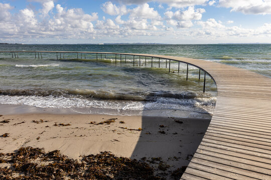 View of The Infinite Bridge extending over the sea with a sandy beach and seaweed in the foreground in Aarhus, Aarhus, Denmark.
