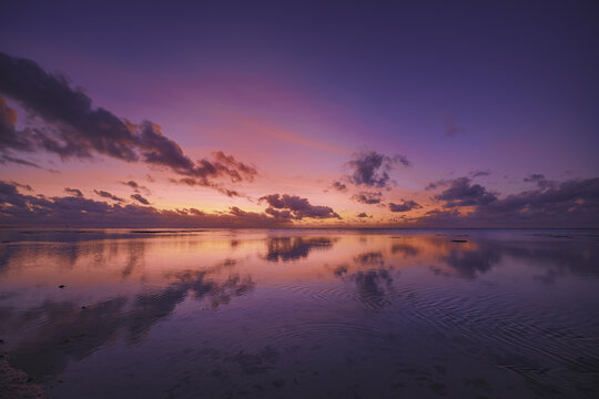 View of Aitutaki Lagoon at sunset with vibrant purple and orange clouds reflecting on the calm water surface in Aitutaki, Cook Islands.