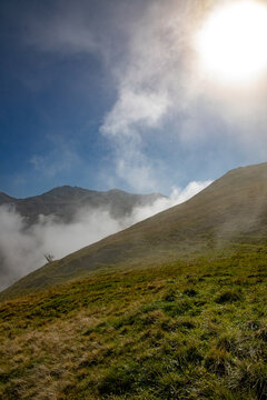 View of the grassy slopes of the Swiss Alps partially covered by low-hanging clouds under a bright sun in Airolo, Ticino, Switzerland.