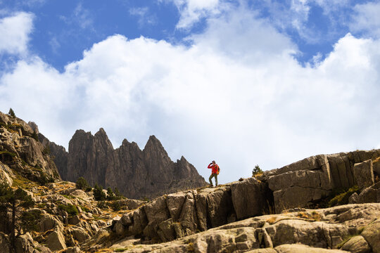 Hiker in red stands on a rocky path. Jagged mountain peaks rise against a bright blue sky. White clouds drift above the scene. The hiker appears calm and purposeful
