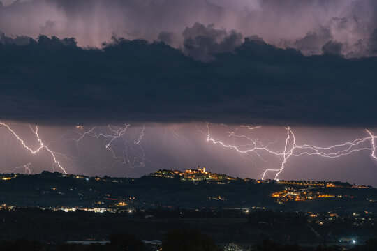 View of the hilltop town of Montecosaro illuminated at night under a dramatic thunderstorm with multiple lightning strikes across the dark sky in Montecosaro, Marche, Italy.