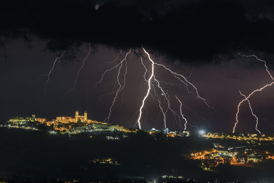 View of multiple lightning bolts striking the night sky over the illuminated historic town of Montecosaro, Marche, Italy.
