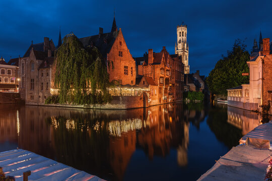 View of the historic Belfry of Bruges illuminated at night, reflecting in the calm canal waters with traditional brick buildings and a large weeping willow tree in Bruges, Flanders, Belgium.