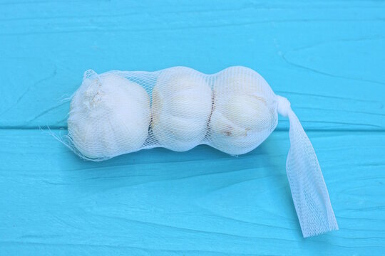 three fresh white heads of garlic in a transparent mesh bag lies on a blue wooden table