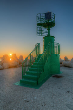 View of a green beacon with metal stairs on a breakwater surrounded by concrete tetrapods during a golden sunrise in Fano, Marche, Italy.