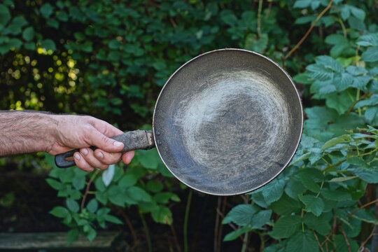 A hand holds a large old gray-black metal frying pan against a backdrop of green vegetation on a summer street