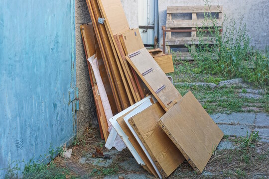 A stack of old brown plywood boards stands against a gray green wall on a street in summer