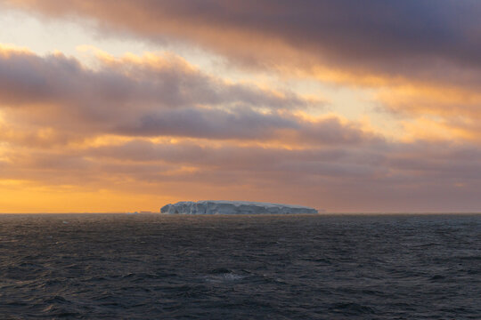 View of an iceberg floating in the ocean under a golden sunset sky in Antarctica.