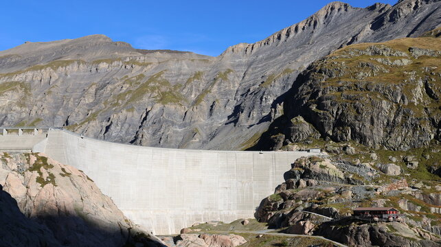 View of the Lac du Vieux Emosson dam nestled against rugged mountain slopes in the Swiss Alps, featuring a concrete wall, rocky terrain, and a small building in Finhaut, Valais, Switzerland.