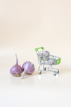 Fresh garlic bulbs with miniature shopping cart for grocery delivery and sale on beige background with copy space