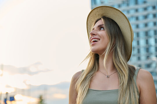 Young woman in straw hat looking up and smiling on city street outdoors