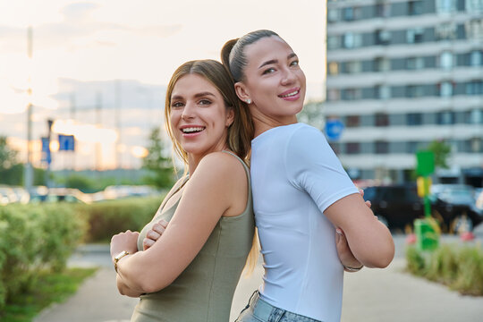 Two smiling young women standing back to back outdoors on city street background
