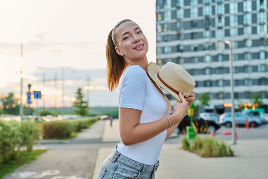 Young woman holding straw hat looking at camera outdoors on city street background