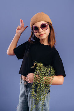 Young girl wearing sunglasses and a beige beanie poses playfully with a plant in her hand against a solid blue background