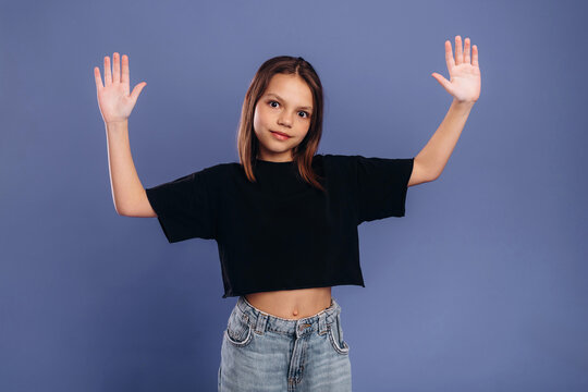 Young girl with brown hair wearing a black cropped t-shirt and blue jeans raises her hands against a solid blue background in a studio setting