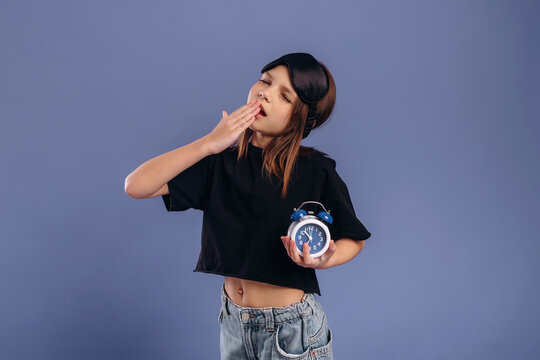 Young girl with long hair yawning while holding a blue alarm clock, wearing a black crop top and denim shorts against a solid purple background