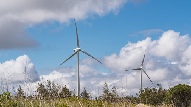 View of wind turbines at Parque Eolico da Serra dos Candeeiros set against a bright blue sky with fluffy white clouds in Rio Maior, Santarem, Portugal.