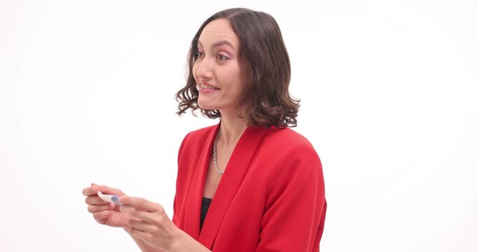 Happy woman in red jacket looks at pregnancy test and jumps up with joy on white backdrop. Young businesswoman excited about results in studio