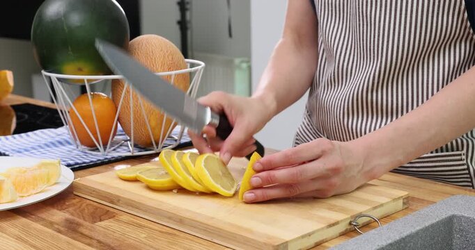 Young woman hands slice lemon with sharp knife on wooden cutting board timelapse. Female in apron cuts sour citrus fruit in modern kitchen