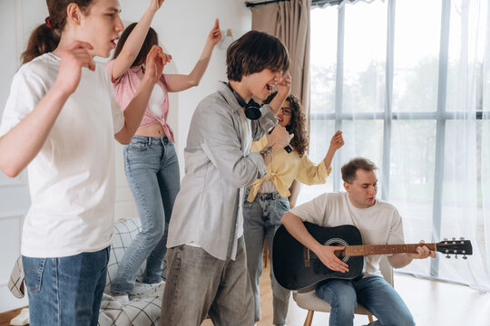 Group of young adults singing and playing guitar in a bright living room with large windows, showcasing a lively indoor atmosphere and casual clothing styles