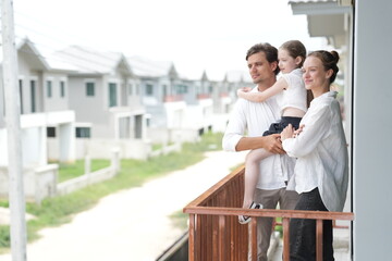 Obraz premium Caucasian family of three embracing look out over the rows of new white houses from the railing standing on the terrace of a newly built suburban home, shallow depth of field