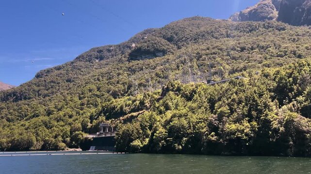 The lakeside works of the Manapouri hydro power station at West Arm, with a wharf, a boom line on the water and transmission gear climbing the bush covered slope. Wide shot